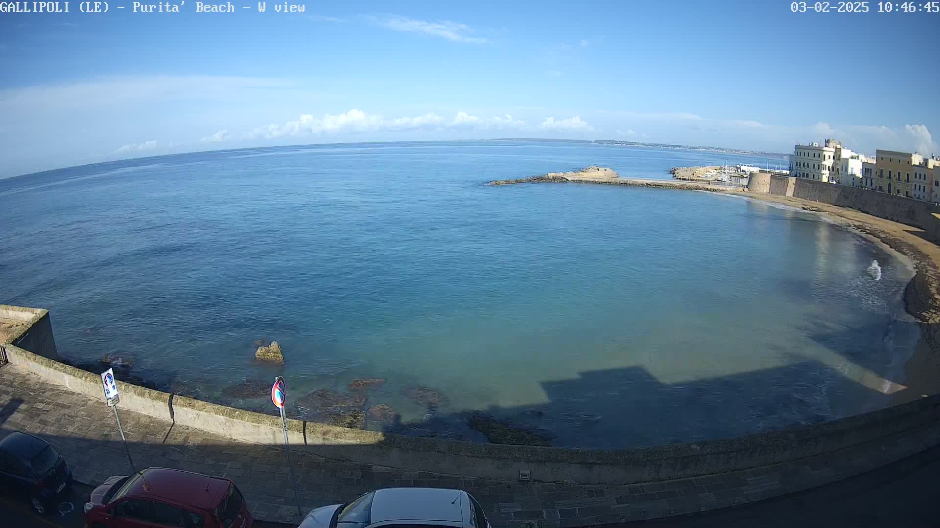 A mostly sunny day reveals a clear blue ocean bay curving along a stone wall and buildings, with a few cars parked on a road nearby.