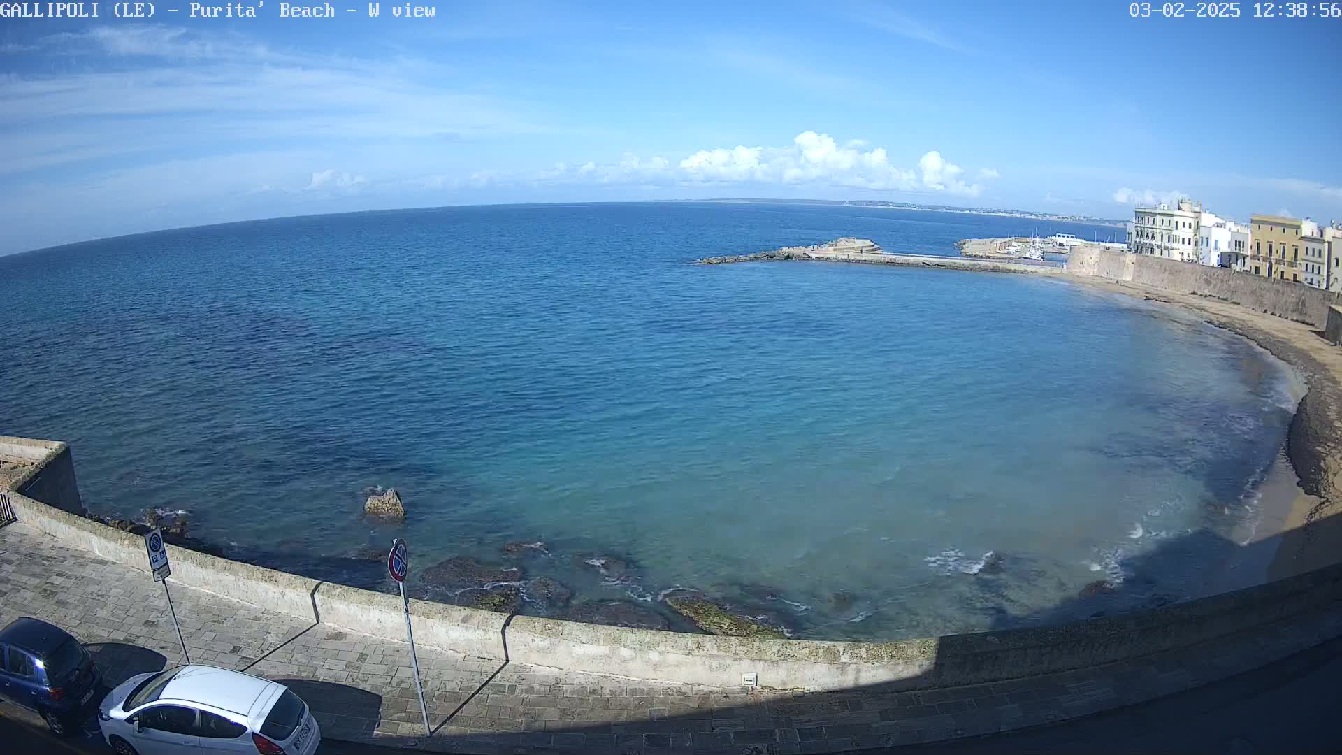 A mostly sunny day shows a calm, clear blue sea curving into a sandy beach next to a stone wall and buildings.