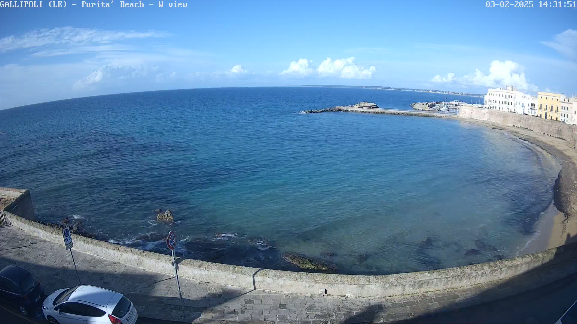 A partly cloudy blue sky looks over a calm, clear turquoise sea curving around a sandy beach and stone buildings.