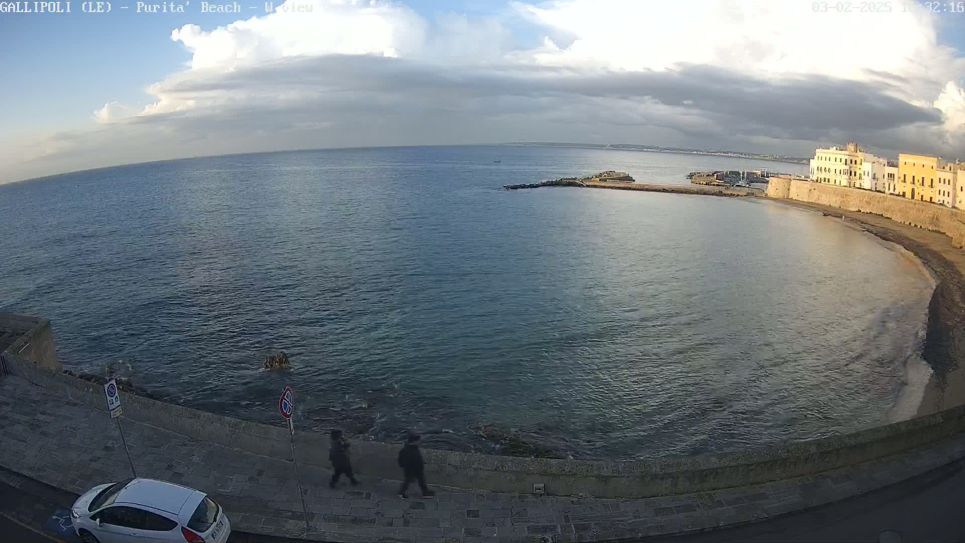 A partly cloudy day overlooks a calm bay with a stone wall and buildings along the shore, where two people walk along a paved path next to a parked car.