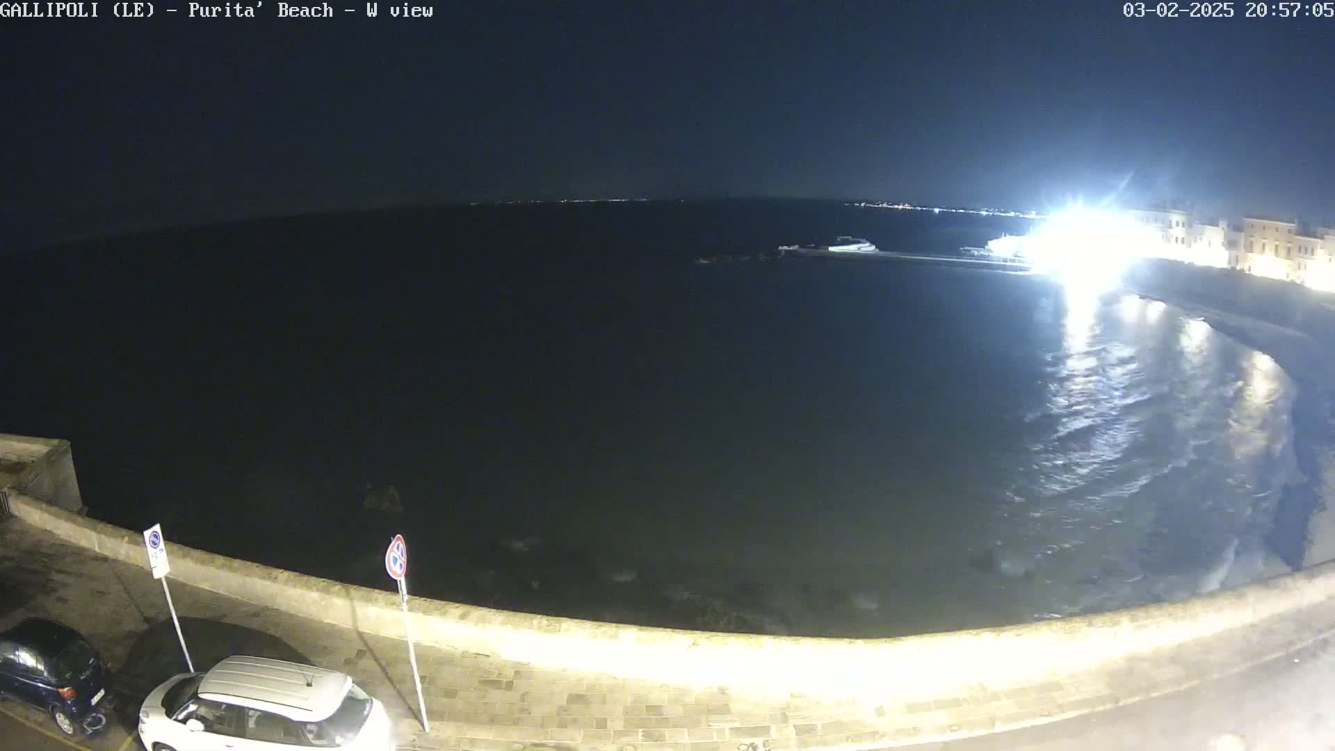 A nighttime view of a calm sea lapping against a stone wall, with a few cars parked along the wall and buildings faintly visible in the distance under a dark sky.