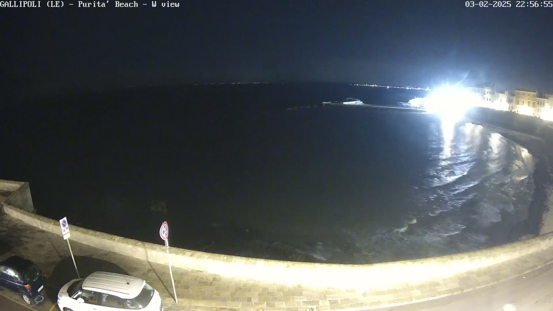 At night, turbulent waves crash against a low stone wall next to a dark body of water, with cars parked along the wall and buildings faintly visible in the distance.