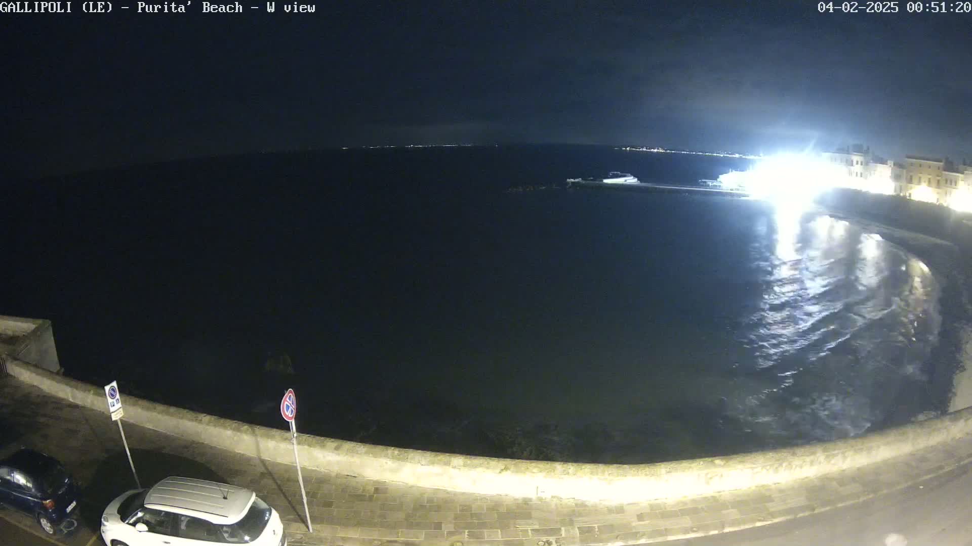 A nighttime, seaside view shows gently lapping waves on a dark, calm sea next to a stone wall and parked cars.
