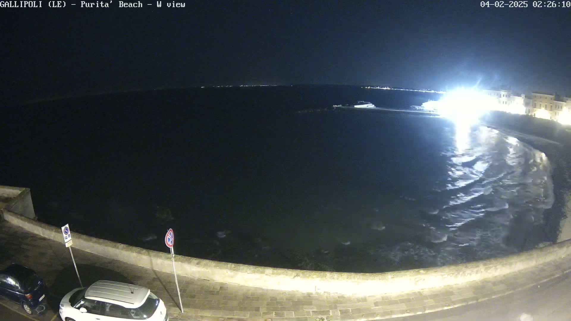 A nighttime view of a dark ocean with small waves lapping against a low stone wall, with a couple of cars parked nearby under artificial light.