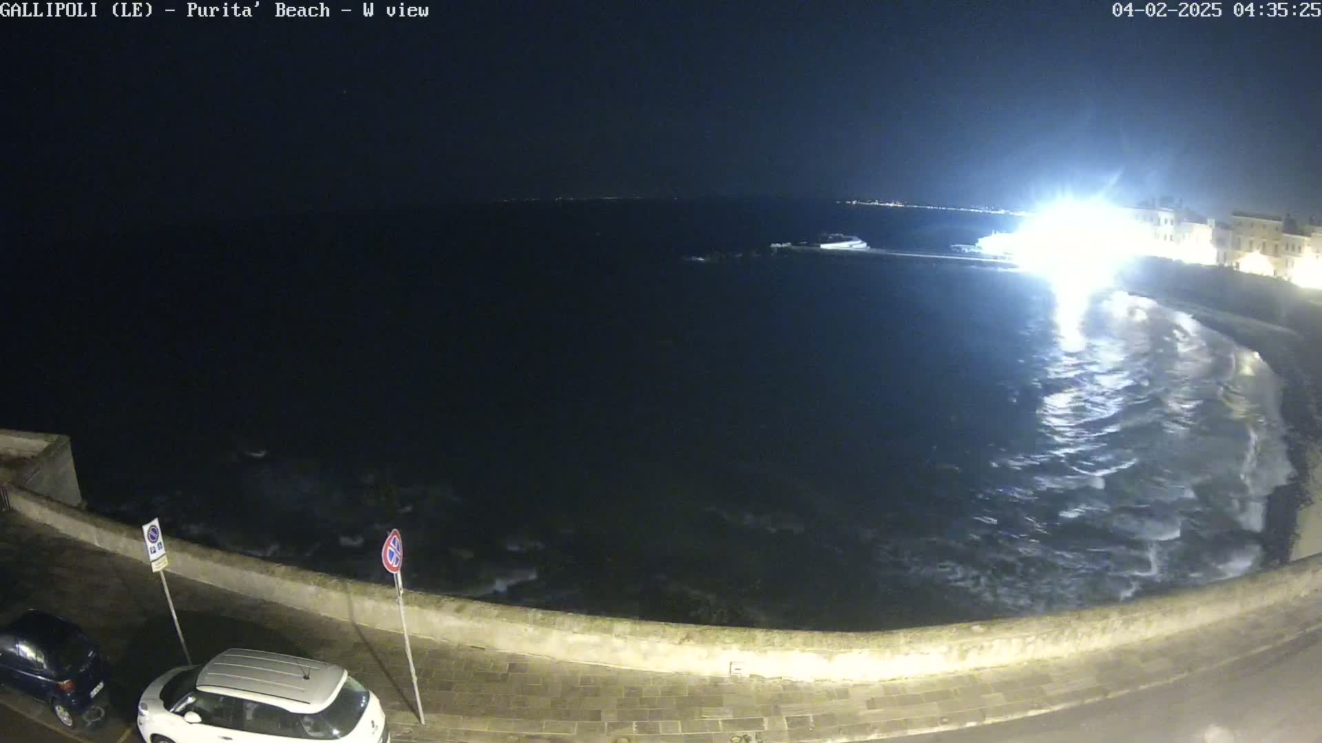A nighttime view of a dark, wavy ocean crashing against a low stone wall next to a street with parked cars under a mostly clear sky.