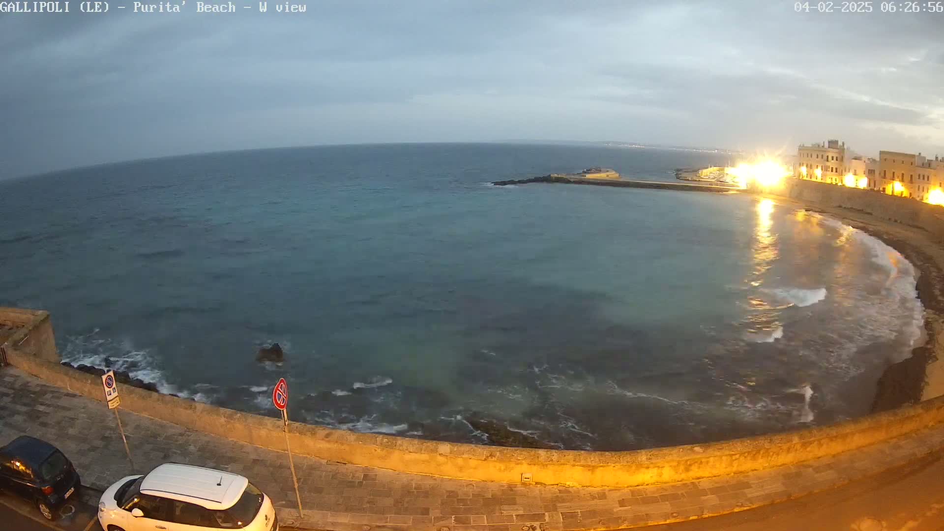 A calm, twilight ocean laps against a stone wall, with a town illuminated in the background.