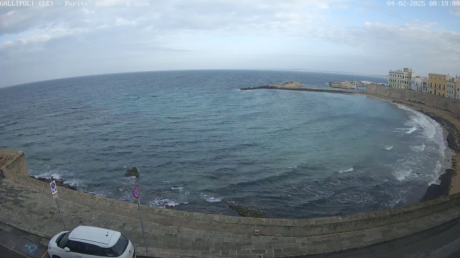 A coastal scene with a partly cloudy sky shows a calm sea lapping a curved shoreline with buildings and a parked car.