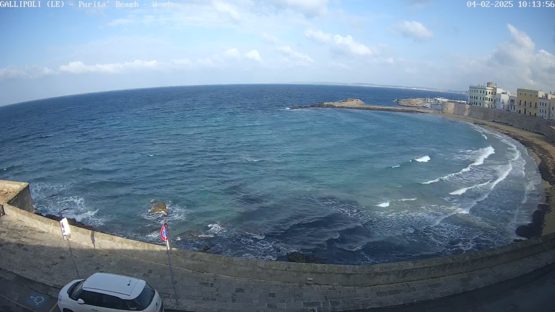 A partly cloudy day shows a coastal scene with a calm sea featuring small waves lapping a stone shore and buildings in the distance.