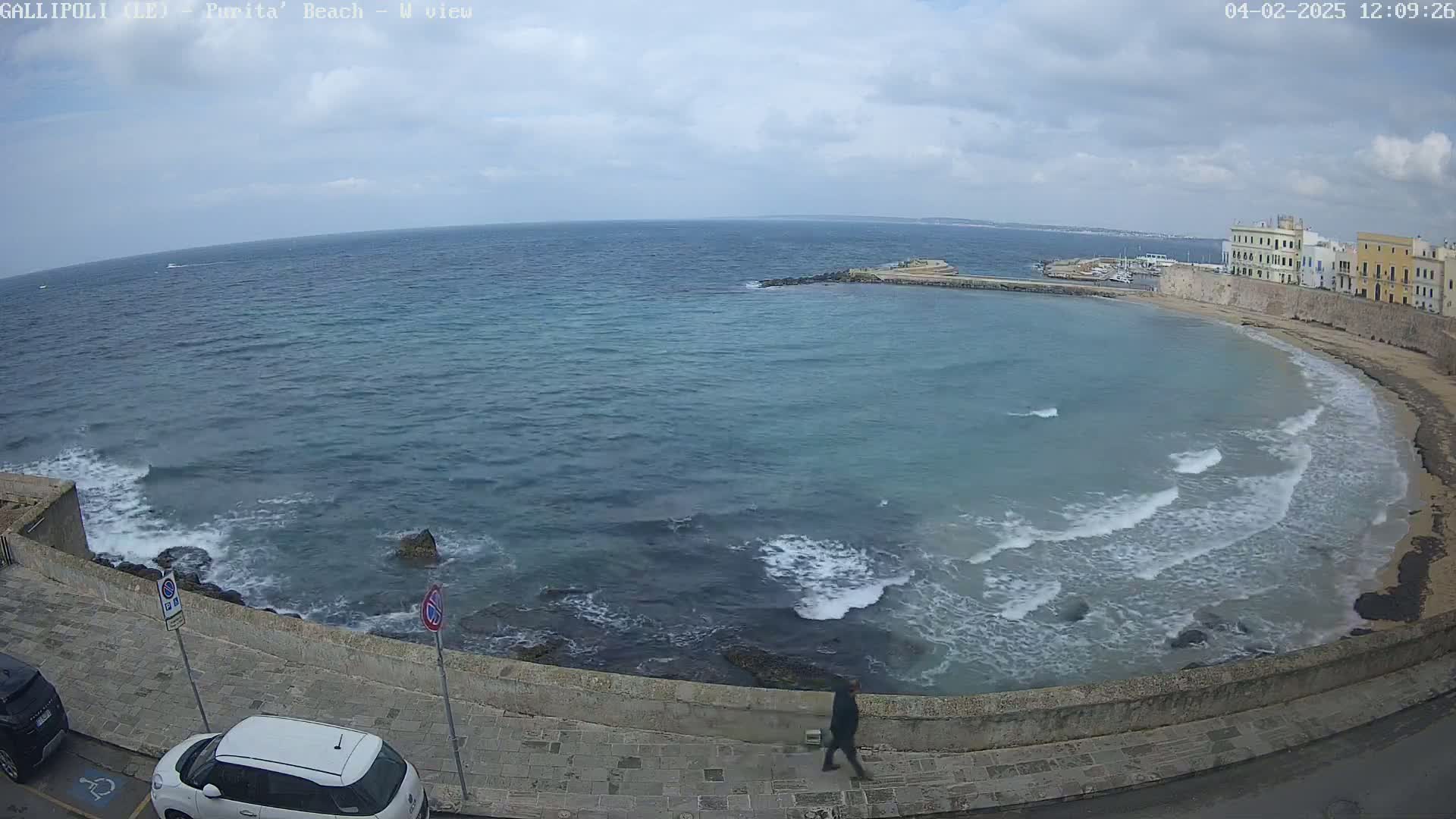 A partly cloudy day shows a view of a calm sea with small waves lapping a sandy beach next to a stone wall and buildings.