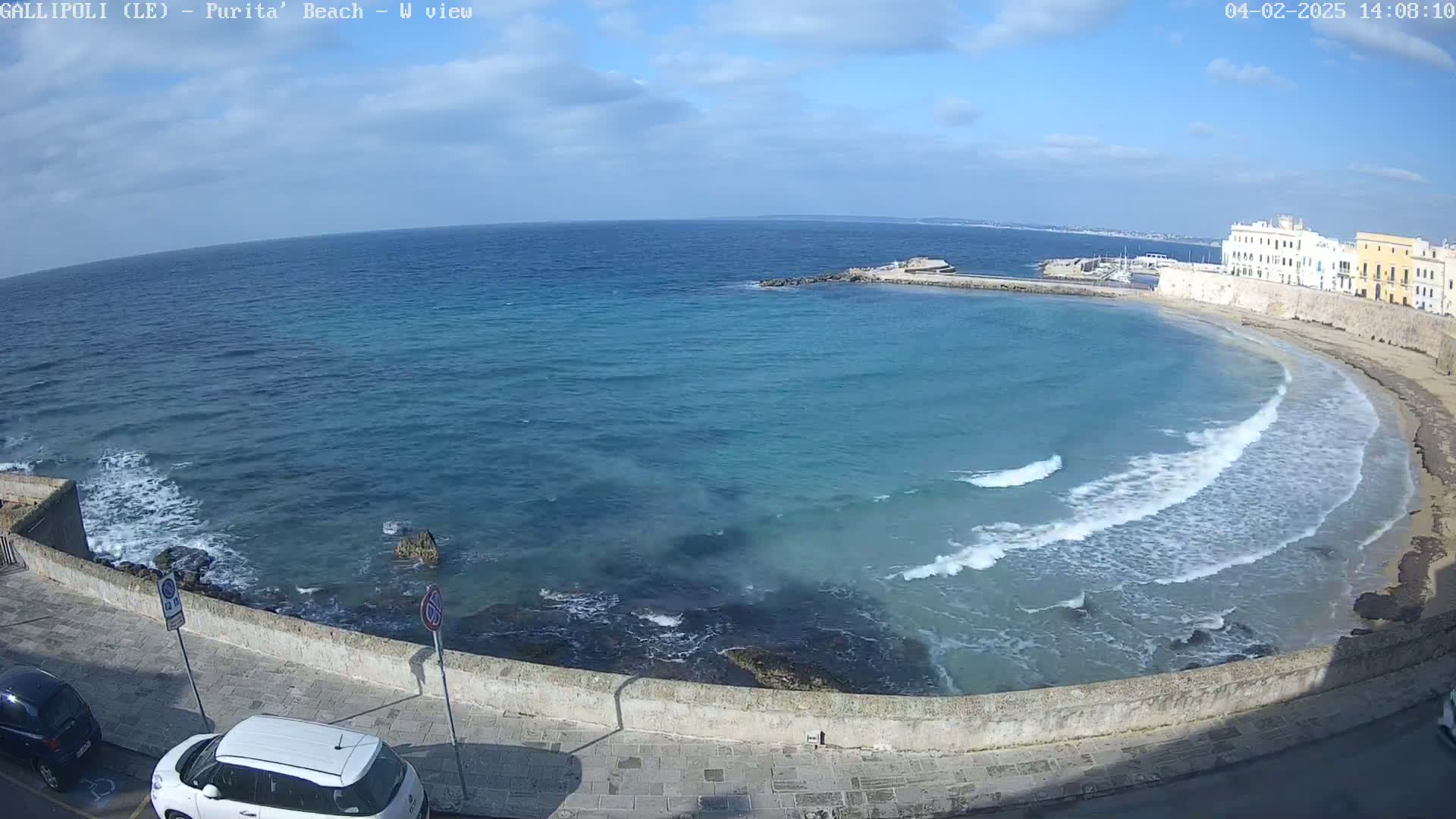 A partly cloudy day shows a view of a tranquil bay with gentle waves lapping a sandy beach, next to a stone wall and buildings.