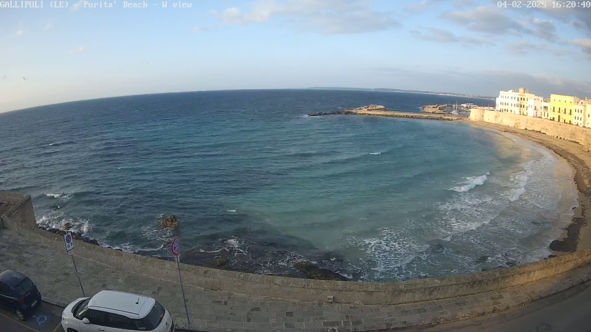 A partly sunny day shows a sandy beach curving around a calm bay with gentle waves lapping a stone wall, and a town in the background.