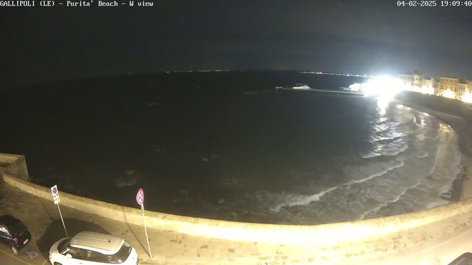A nighttime view of a dark, wavy ocean lapping against a shoreline with a low wall and parked cars, under a clear sky.