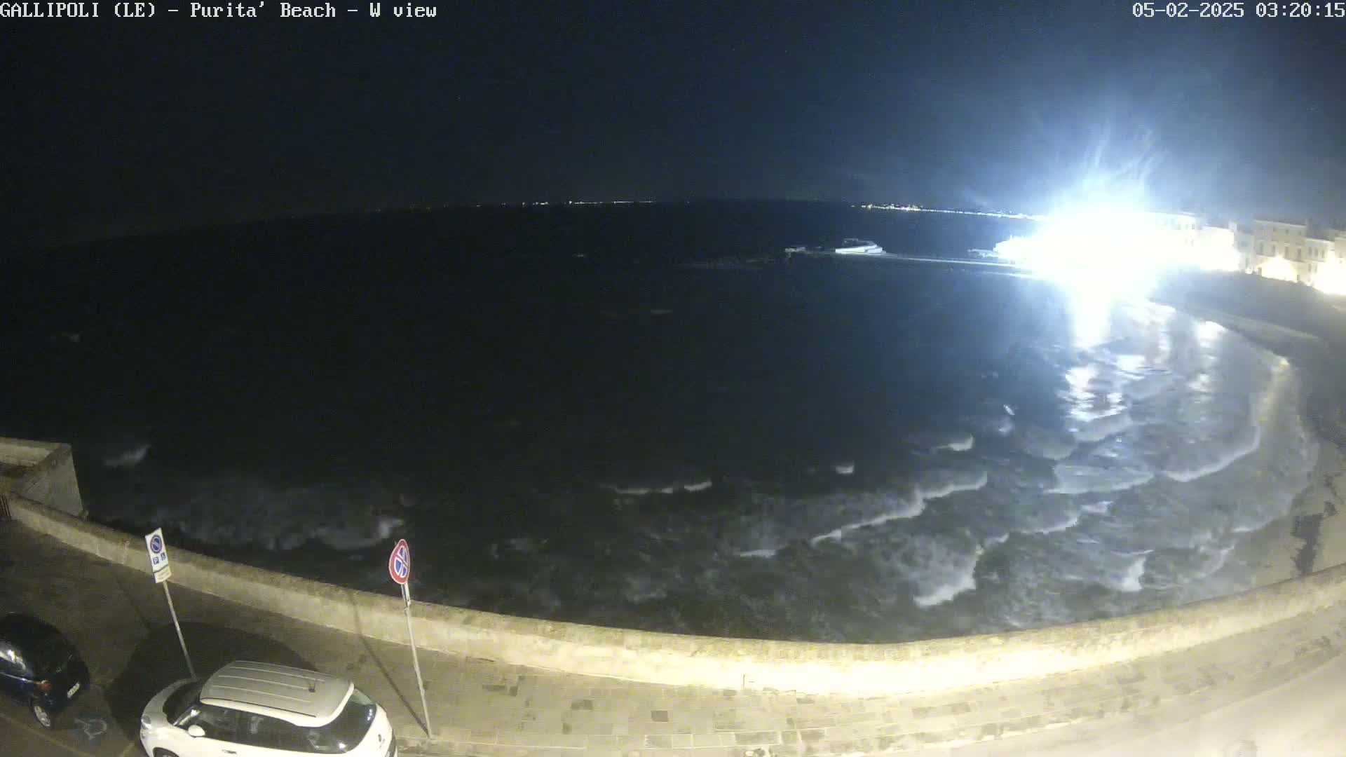 A nighttime view of a wavy sea lapping against a low stone wall, with cars parked along a paved road beside it.
