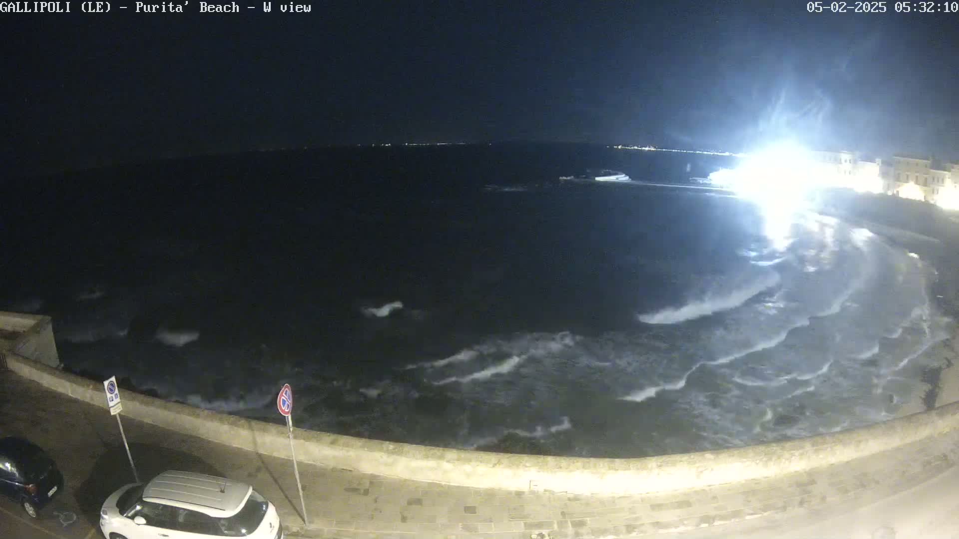 A nighttime view of a wavy sea crashing against a low stone wall next to a road with parked cars under a dark sky.
