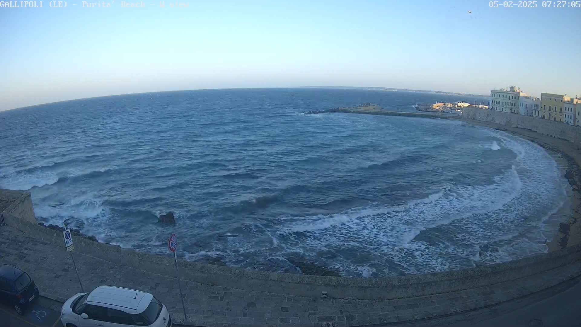 A wavy sea laps against a stone wall bordering a town under a clear sky.