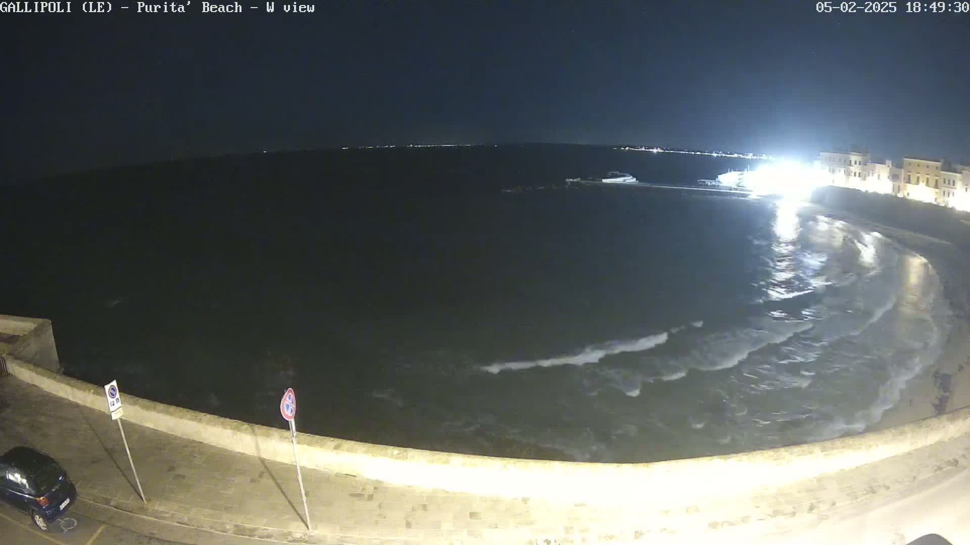 At night, a dark body of water with small waves laps against a low stone wall next to a street with a car parked, under a clear night sky.