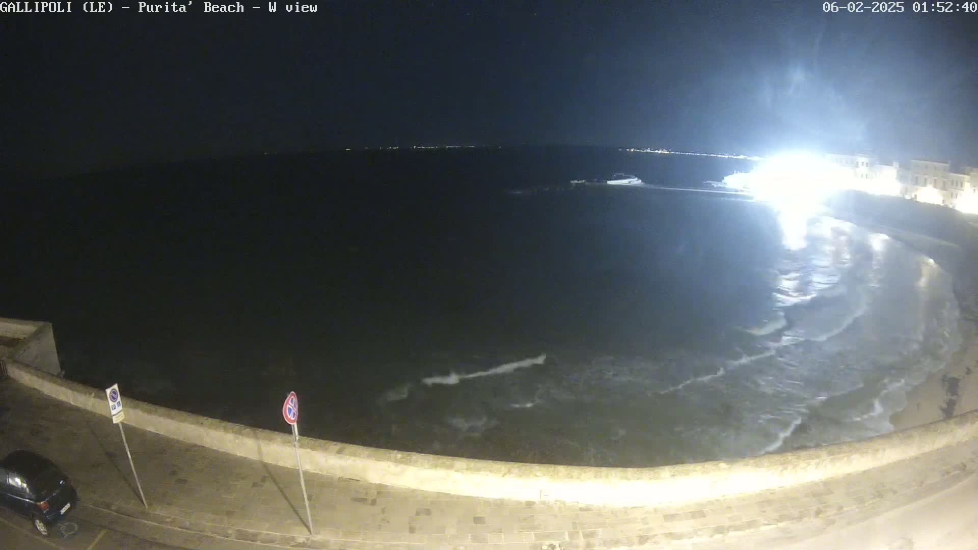 A nighttime view of a dark ocean with small waves lapping a shoreline, a stone wall, and a car parked nearby, under a clear, starlit sky.