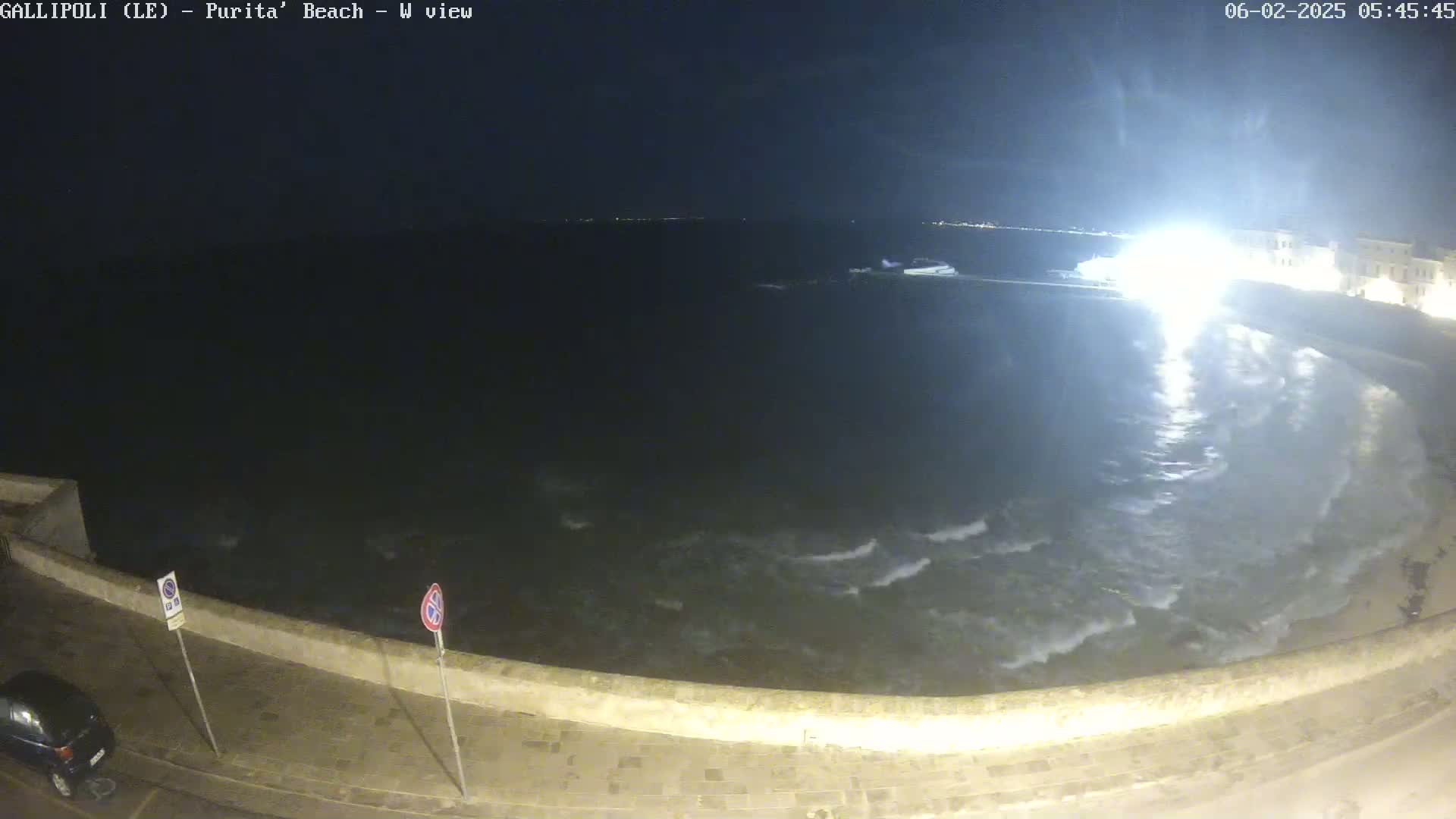 A nighttime view of a wavy ocean lapping against a stone wall, with a car parked nearby under somewhat bright light.