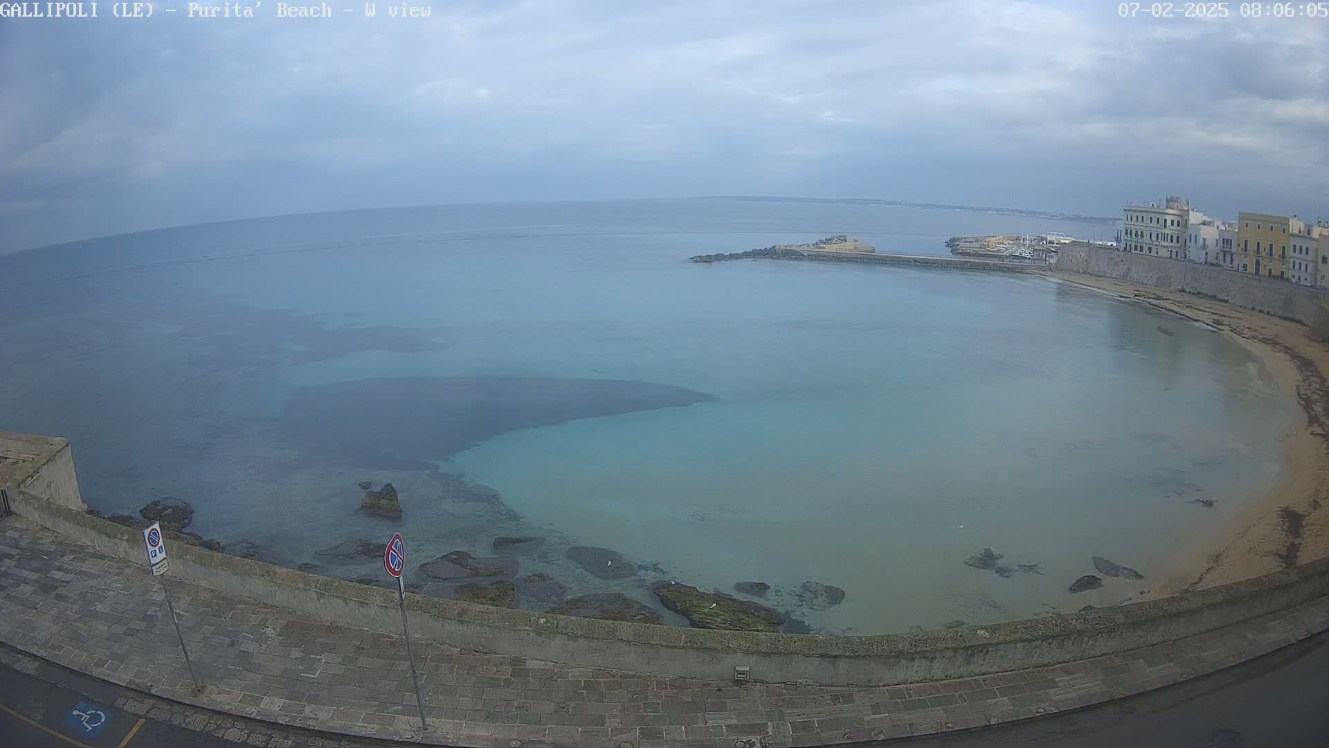 A coastal town's stone wall overlooks a calm, light-blue bay under an overcast sky.