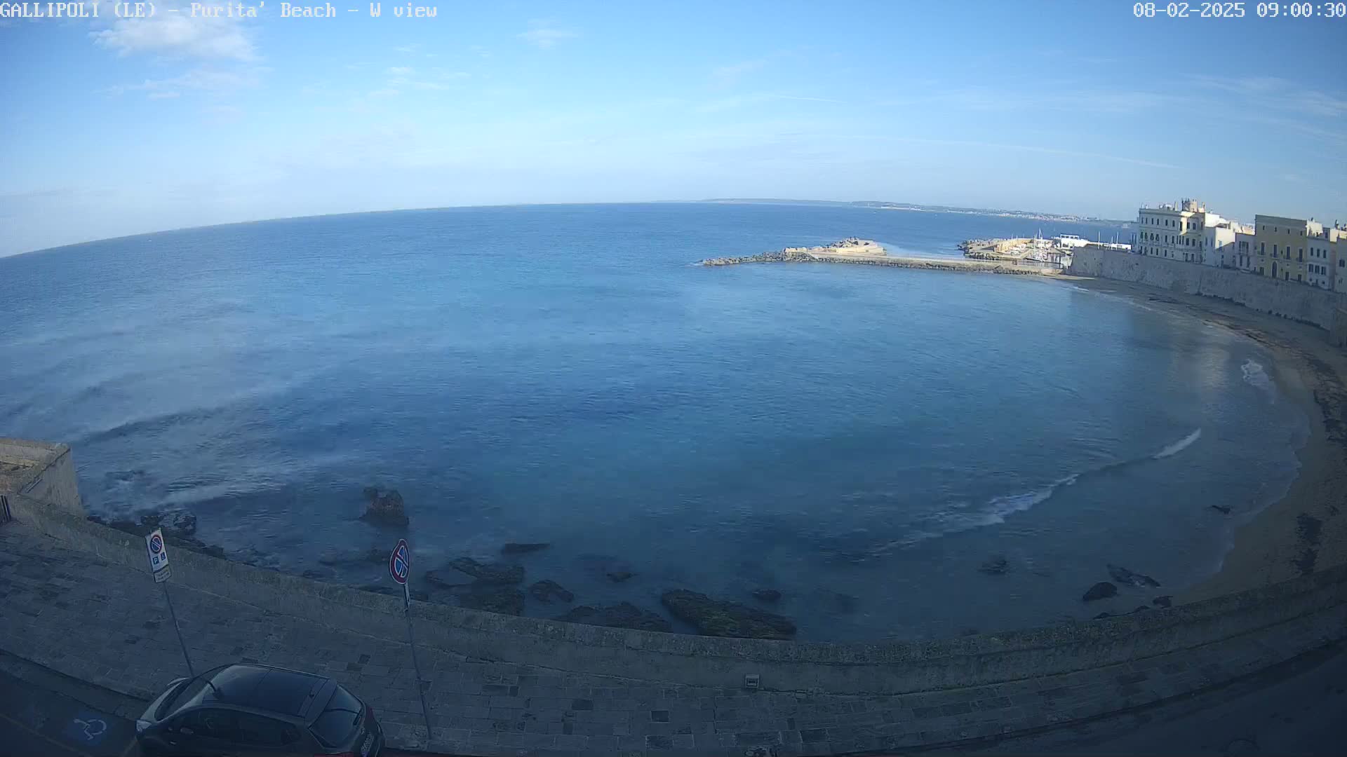 A clear, sunny day overlooks a calm, blue sea curving into a sandy beach next to a stone-walled city.