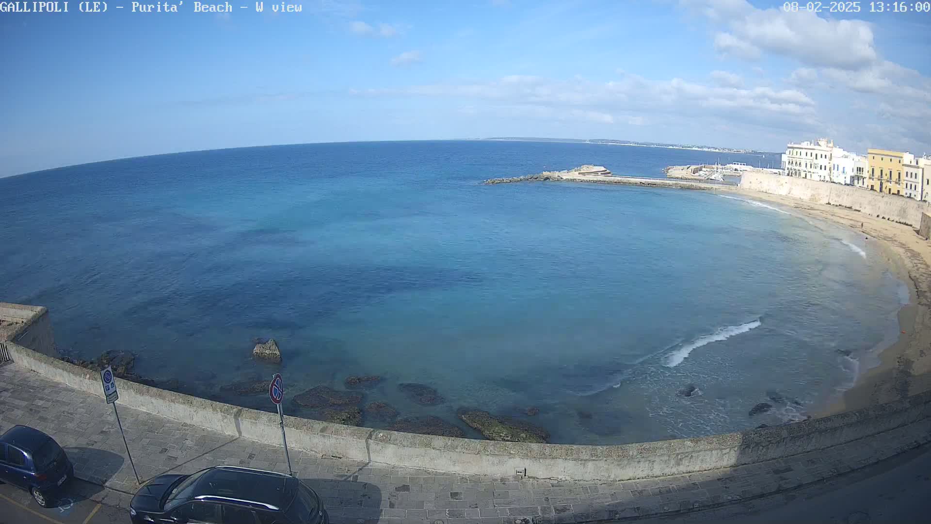 A partly sunny day shows a calm sea curving around a sandy beach next to a stone wall and buildings, with two cars parked nearby.