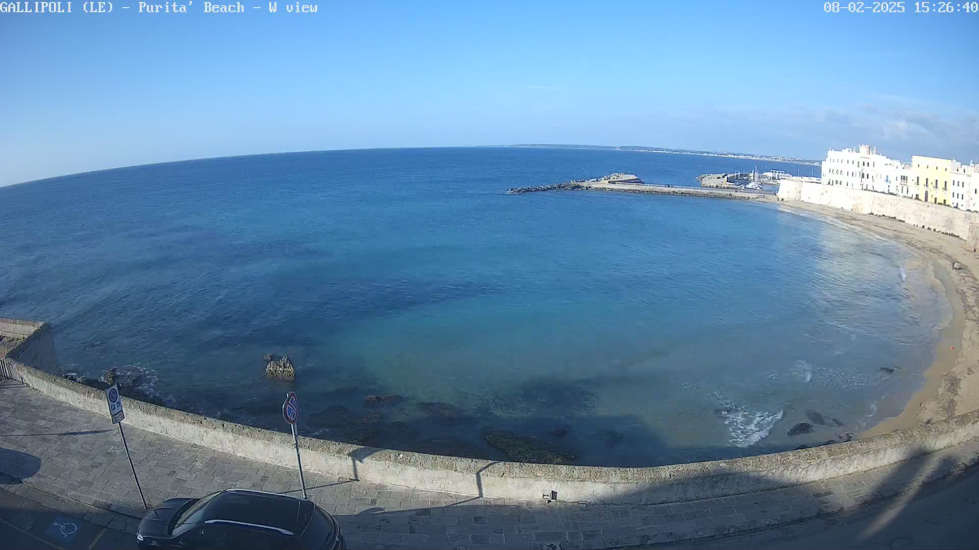 A clear, sunny day overlooking a calm, blue sea, a sandy beach, and a stone wall bordering a paved road with a parked car.