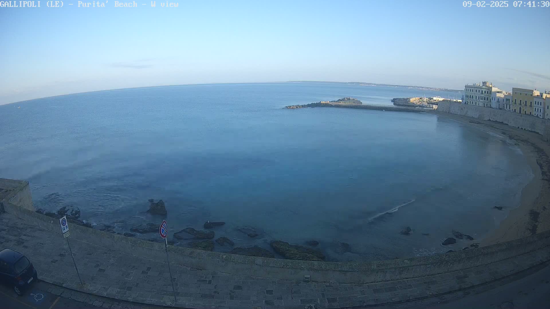 A calm, clear blue sea laps a sandy beach next to a stone wall and buildings under a bright, clear sky.