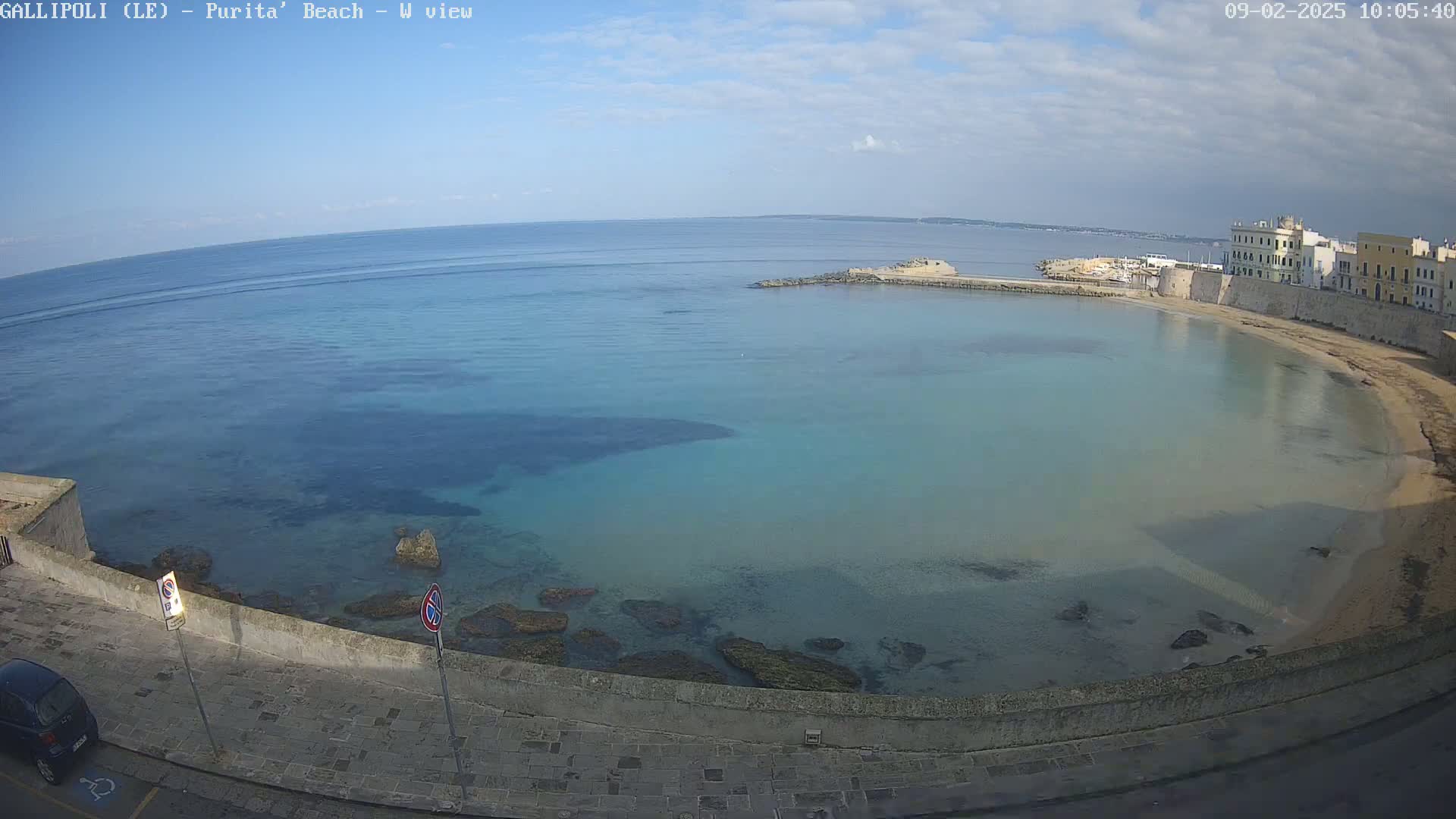A mostly sunny day shows a calm, shallow, teal-colored bay curving around a sandy beach next to a stone wall and buildings.