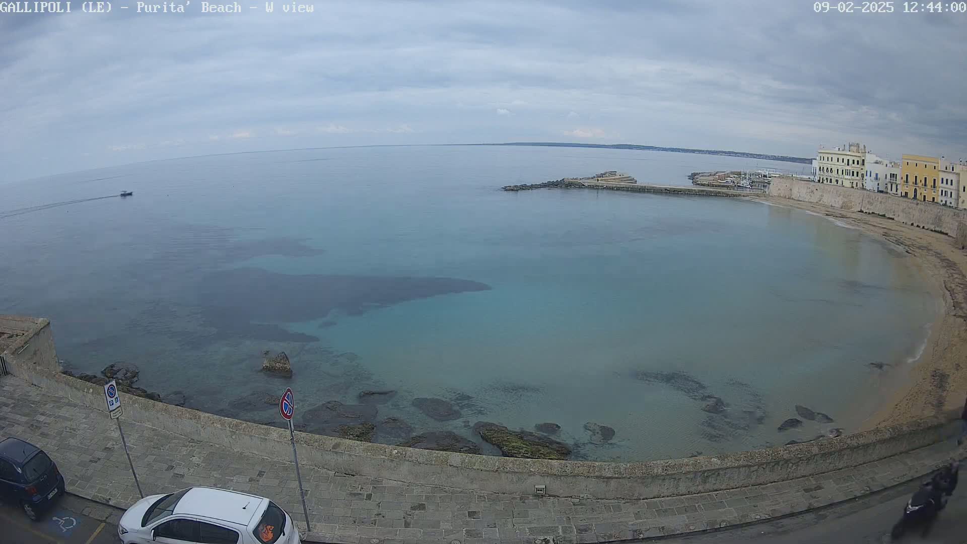 A partly cloudy day reveals a calm, teal-colored bay with a sandy beach and buildings along the shoreline.