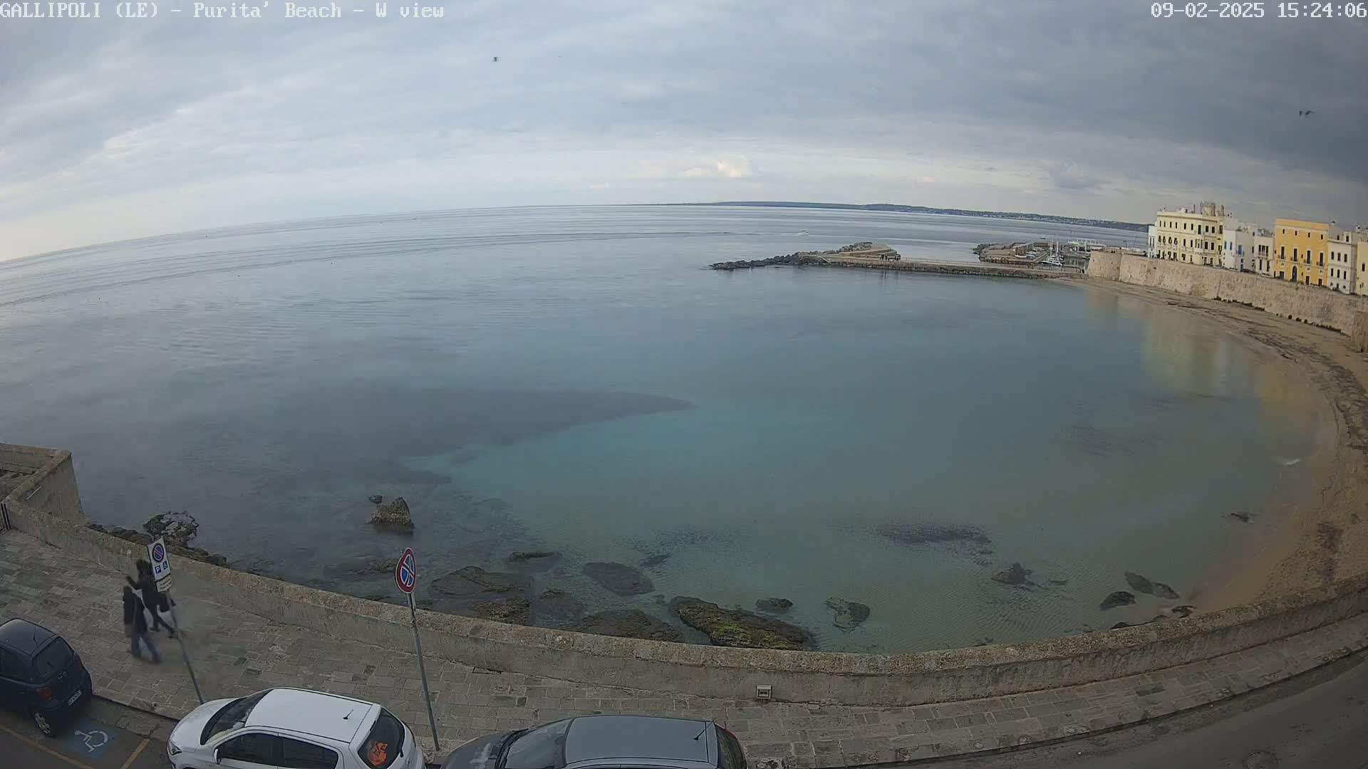 A partly cloudy day shows a calm, teal-colored bay next to a stone walkway and buildings along a sandy beach.
