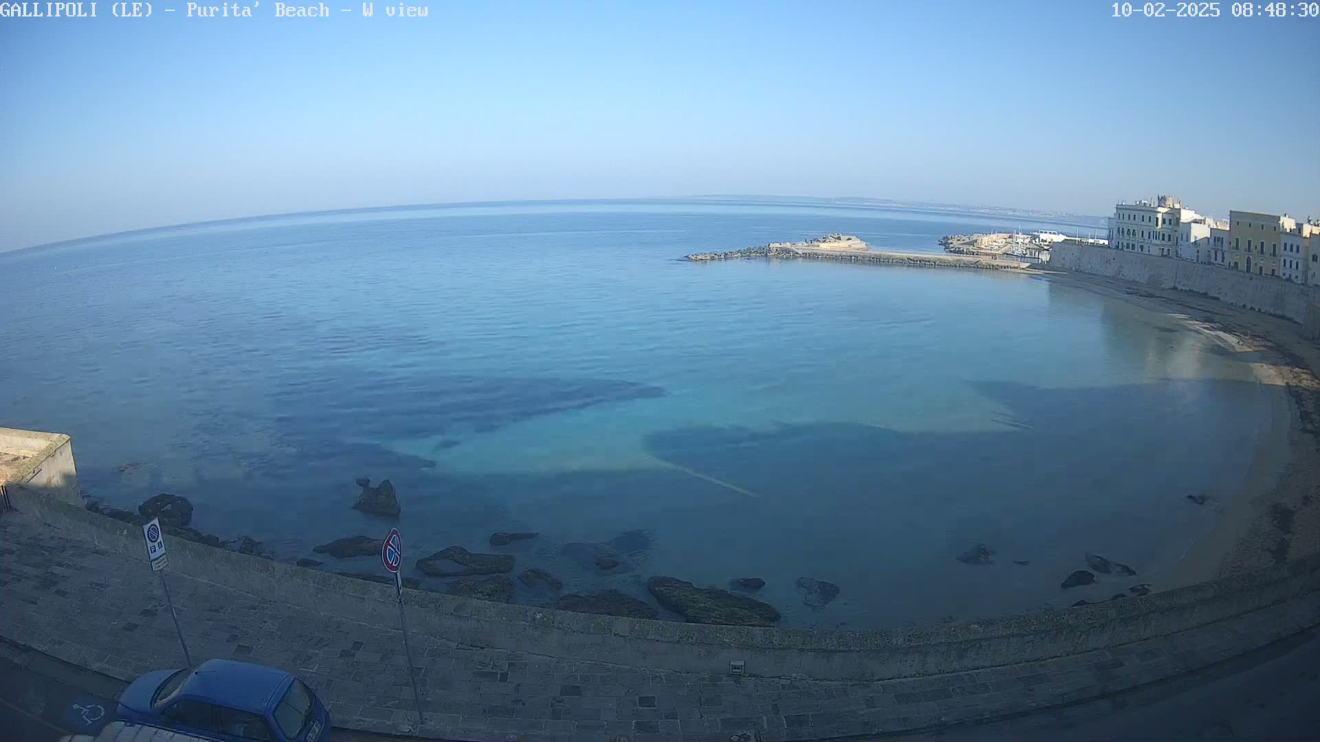A clear, sunny day overlooks a calm, teal-colored ocean bay next to a stone shoreline and buildings.