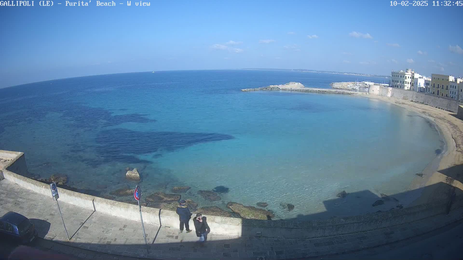 A clear, sunny day reveals a tranquil turquoise bay with a sandy beach, bordered by a low stone wall and buildings, and two people standing near the wall.