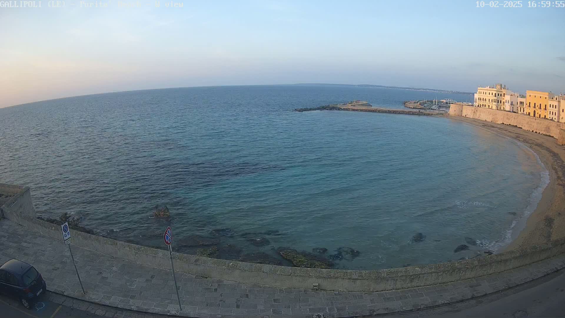 A calm, clear-blue sea laps a sandy beach curving around a stone waterfront lined with buildings under a pale-blue sky.