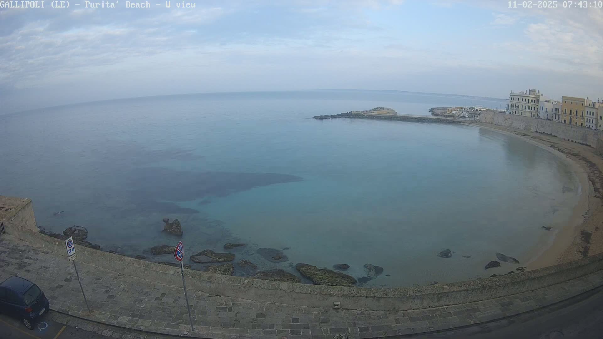 A partly sunny, calm morning scene overlooking a curved bay with clear, shallow turquoise water, rocky shores, a sandy beach, and buildings along the coastline.