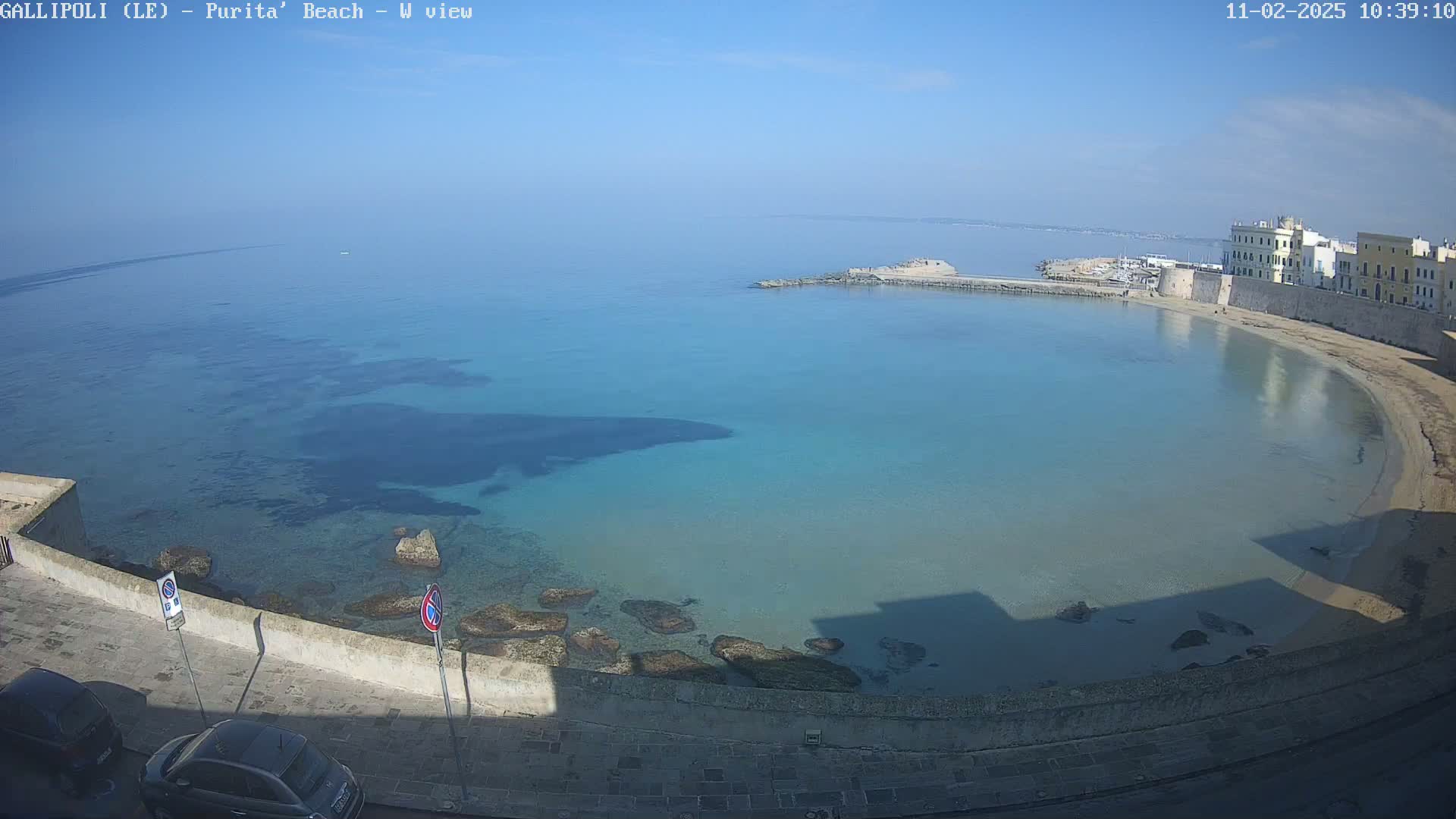 A clear, sunny day reveals a calm, turquoise bay with a sandy beach curving alongside a stone wall and buildings.