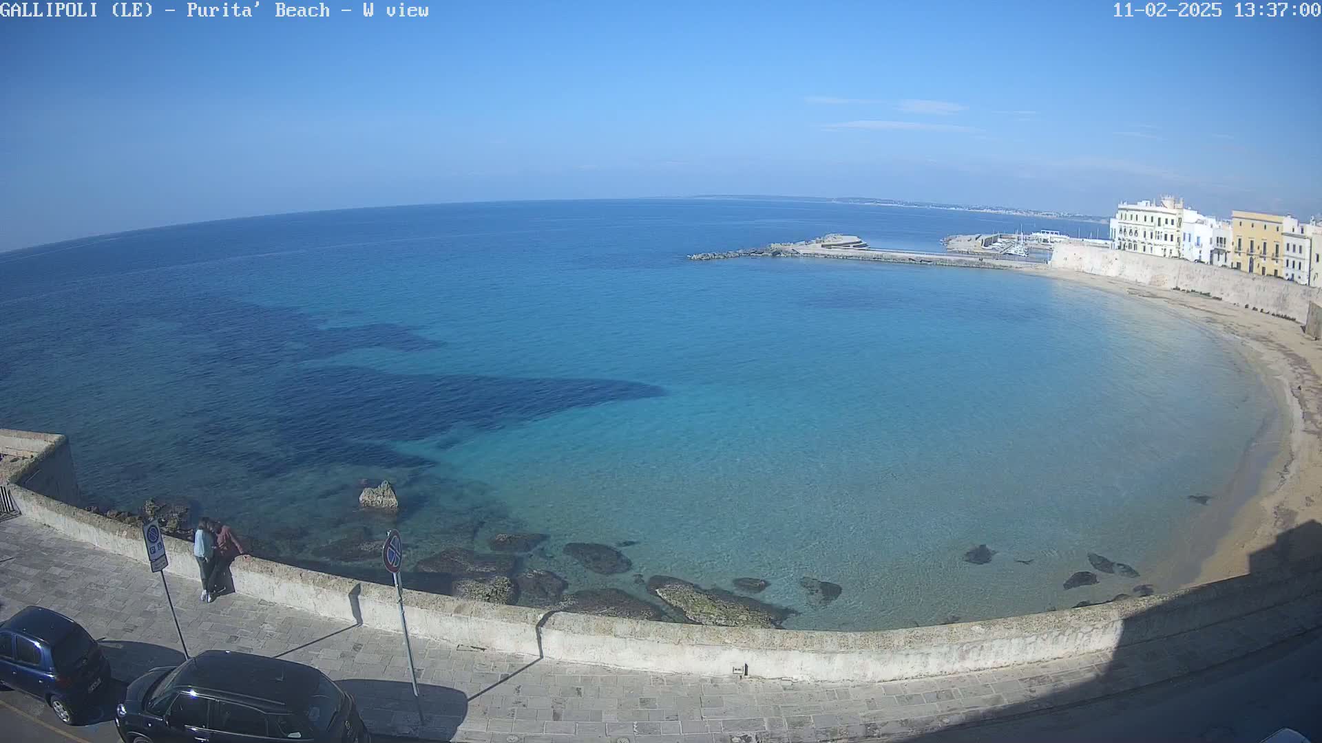 A clear, sunny day reveals a calm, turquoise bay with a sandy beach and buildings along the shoreline.