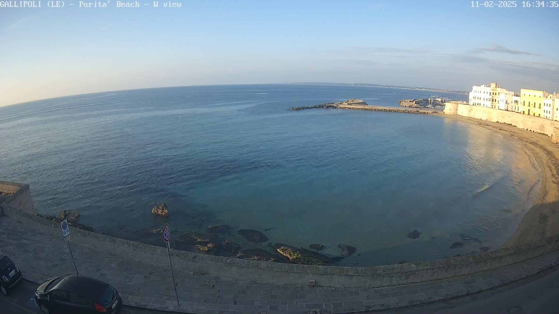 A clear, sunny day overlooks a calm, turquoise bay with a sandy beach and stone buildings along the shoreline.