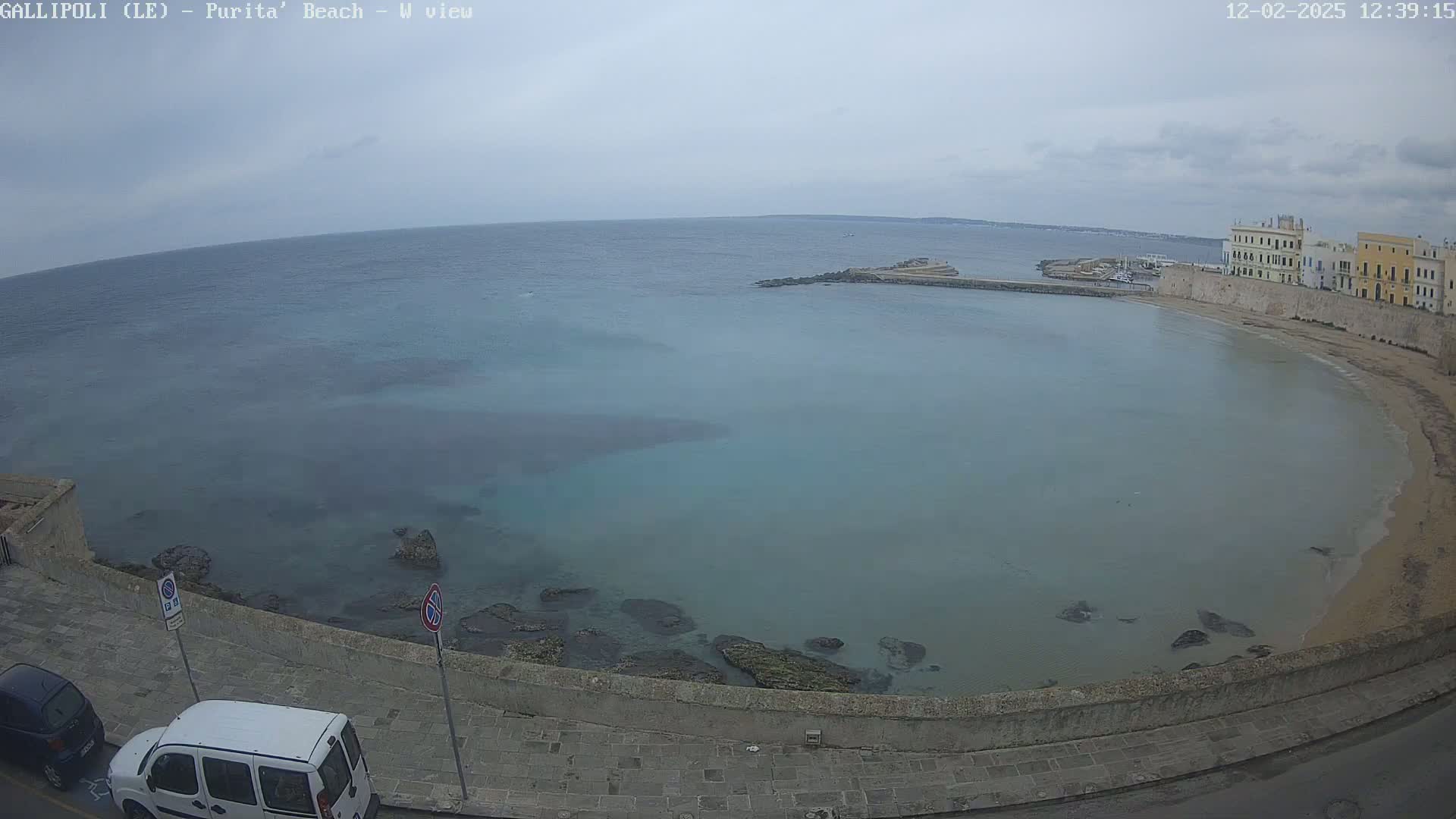 A partly cloudy day overlooks a calm, light-blue bay curving around a sandy beach and stone buildings.