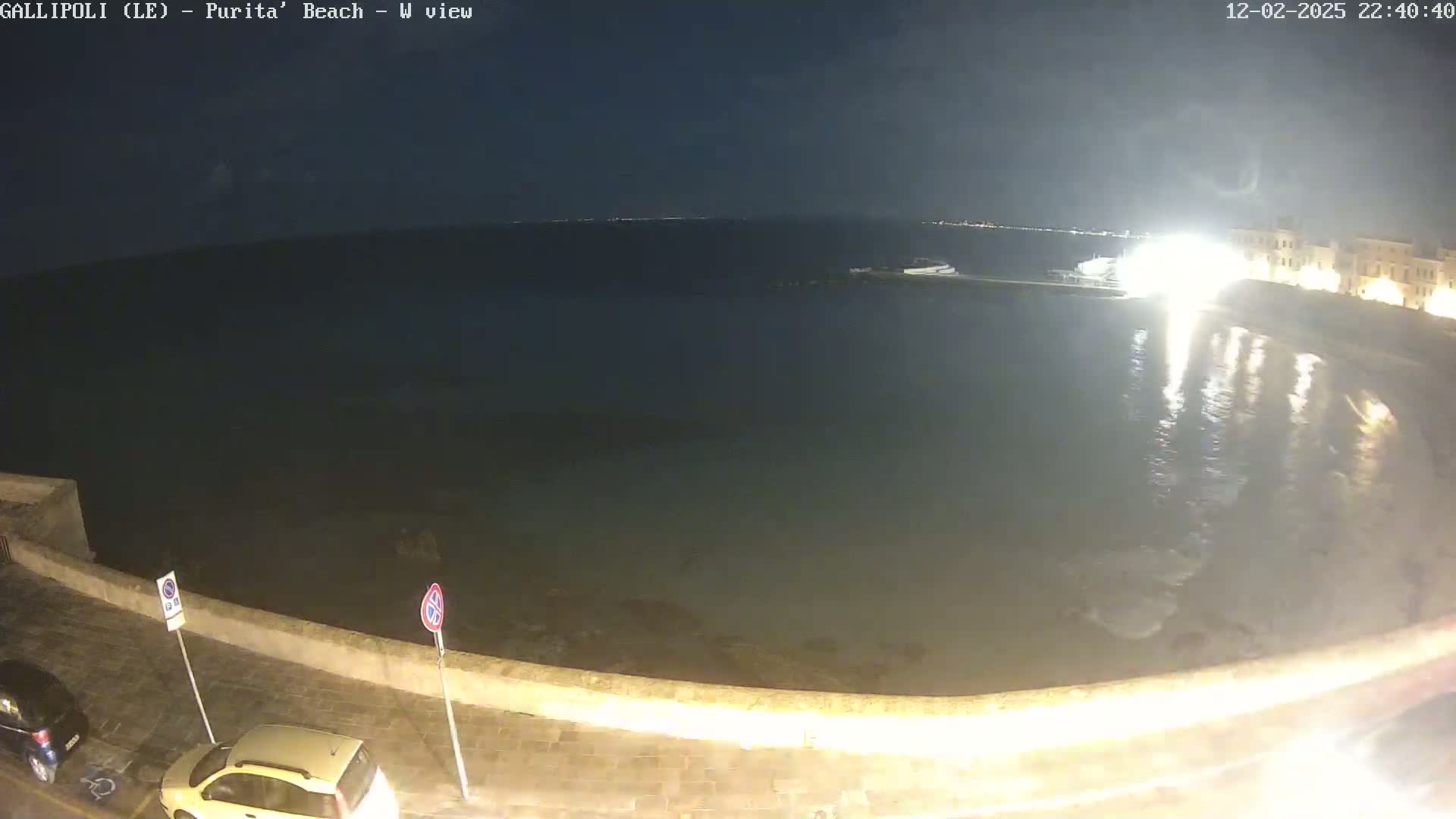 A nighttime view of a calm body of water next to a stone wall and parked cars under a dark sky.