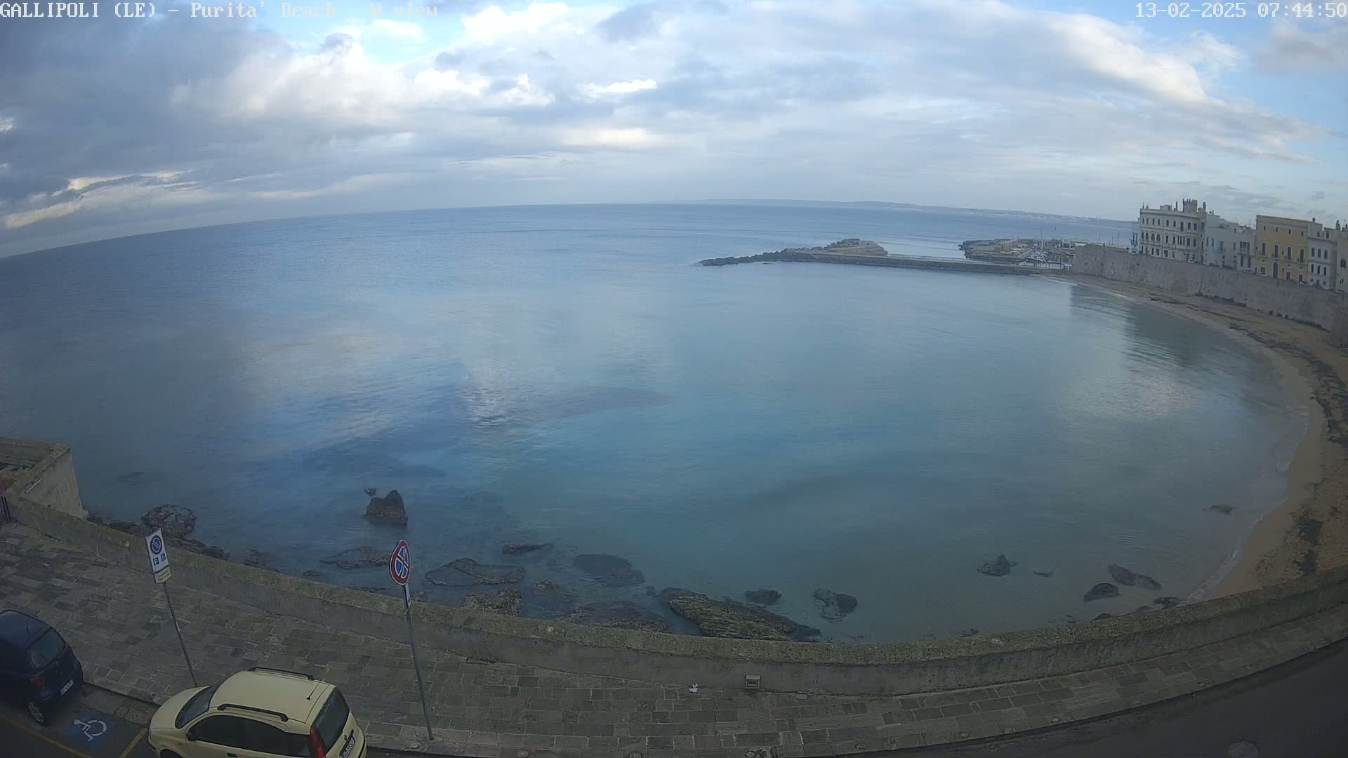 A partly cloudy day overlooks a calm bay with a sandy beach and buildings along the shoreline.