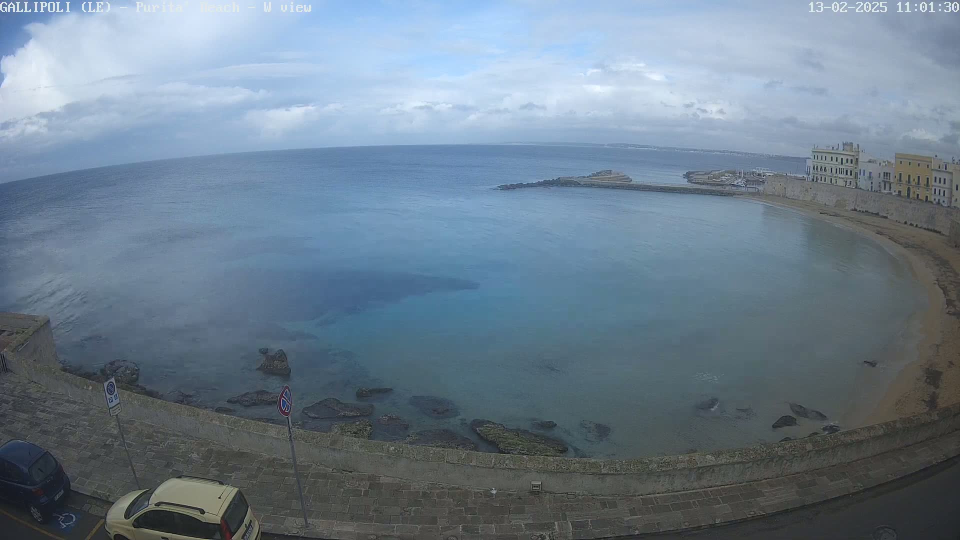 A partly cloudy day overlooks a calm, teal-colored bay with a sandy beach and buildings along the shoreline.