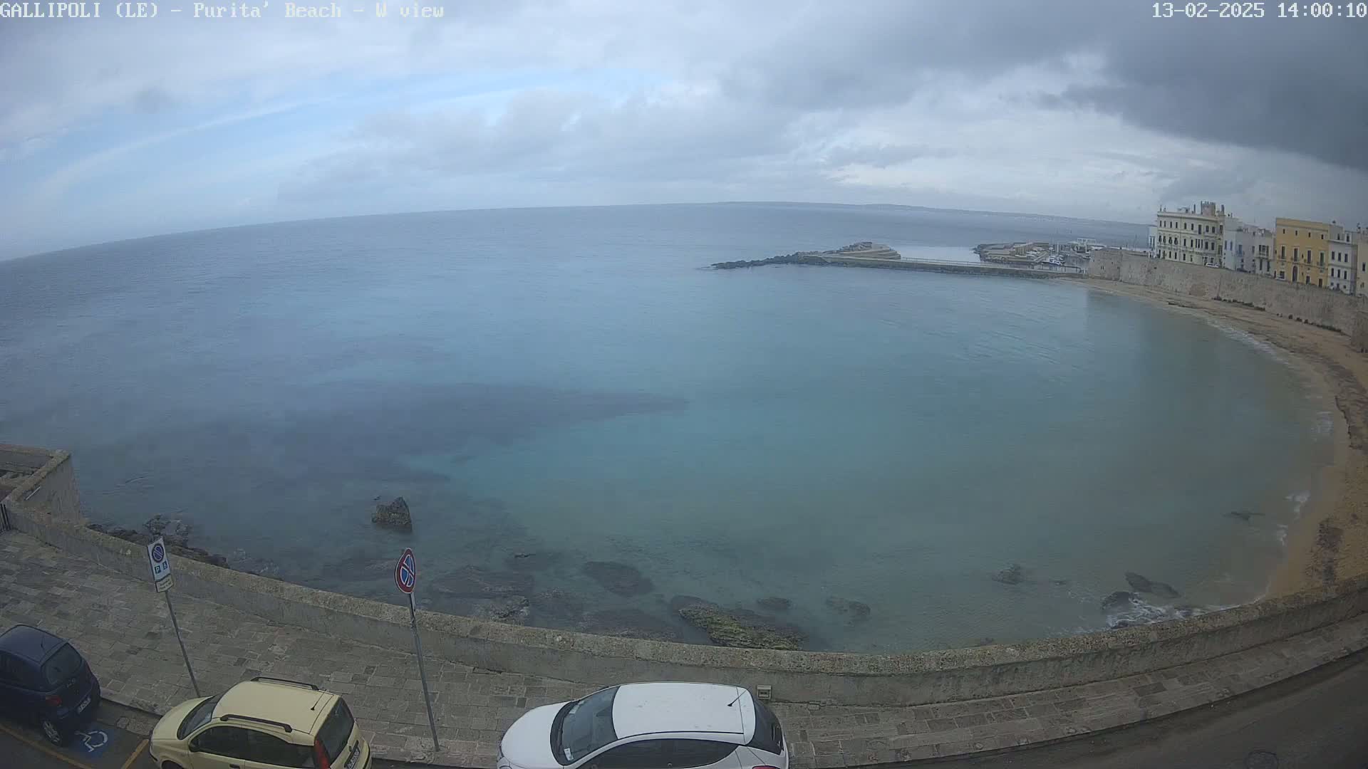 A calm, light-blue bay curves around a stone wall next to a town under an overcast sky.