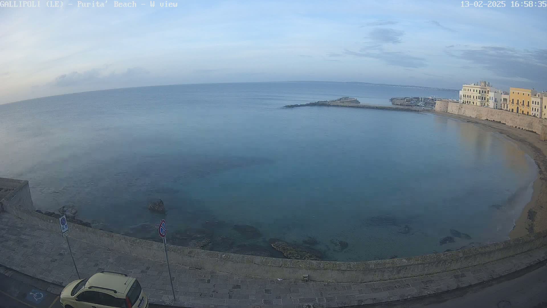 A calm, light-blue sea laps a sandy beach next to a stone wall and buildings under a mostly clear sky.