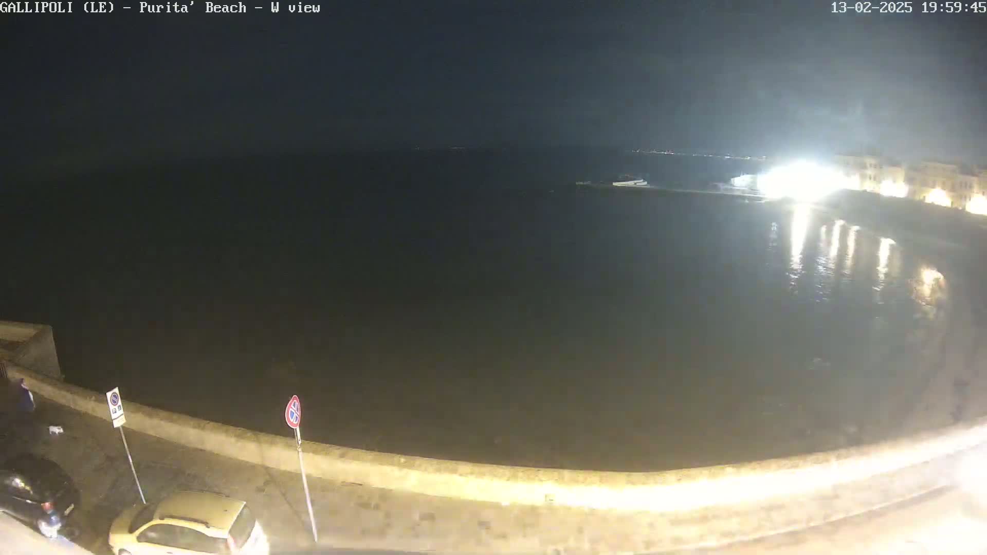 A nighttime view of a calm, dark body of water next to a stone wall with parked cars and a building with lights visible on the right.