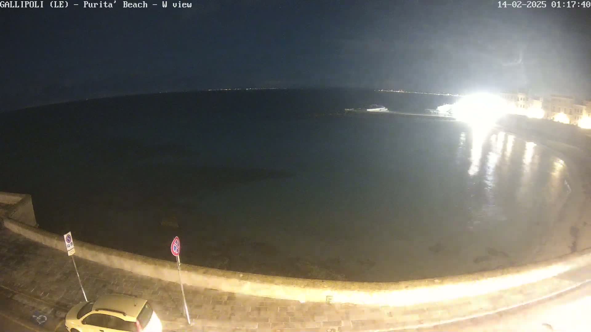 A nighttime view of a calm, dark body of water next to a stone wall and road, with a single car parked nearby under a clear, dark sky.
