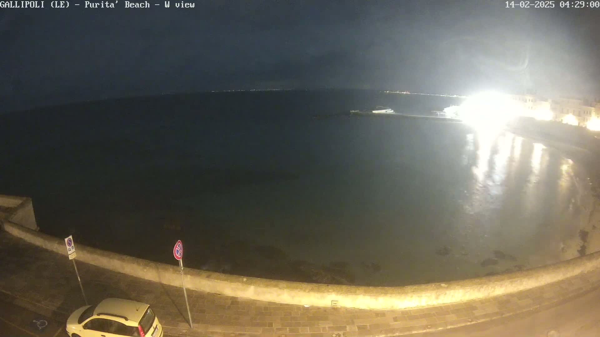 A nighttime view of a calm body of water next to a stone wall, with a car parked on the street and bright lights reflecting on the water.