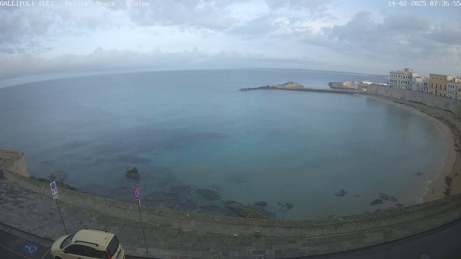 A partly cloudy day overlooks a calm, teal bay curving around a sandy beach and stone buildings.
