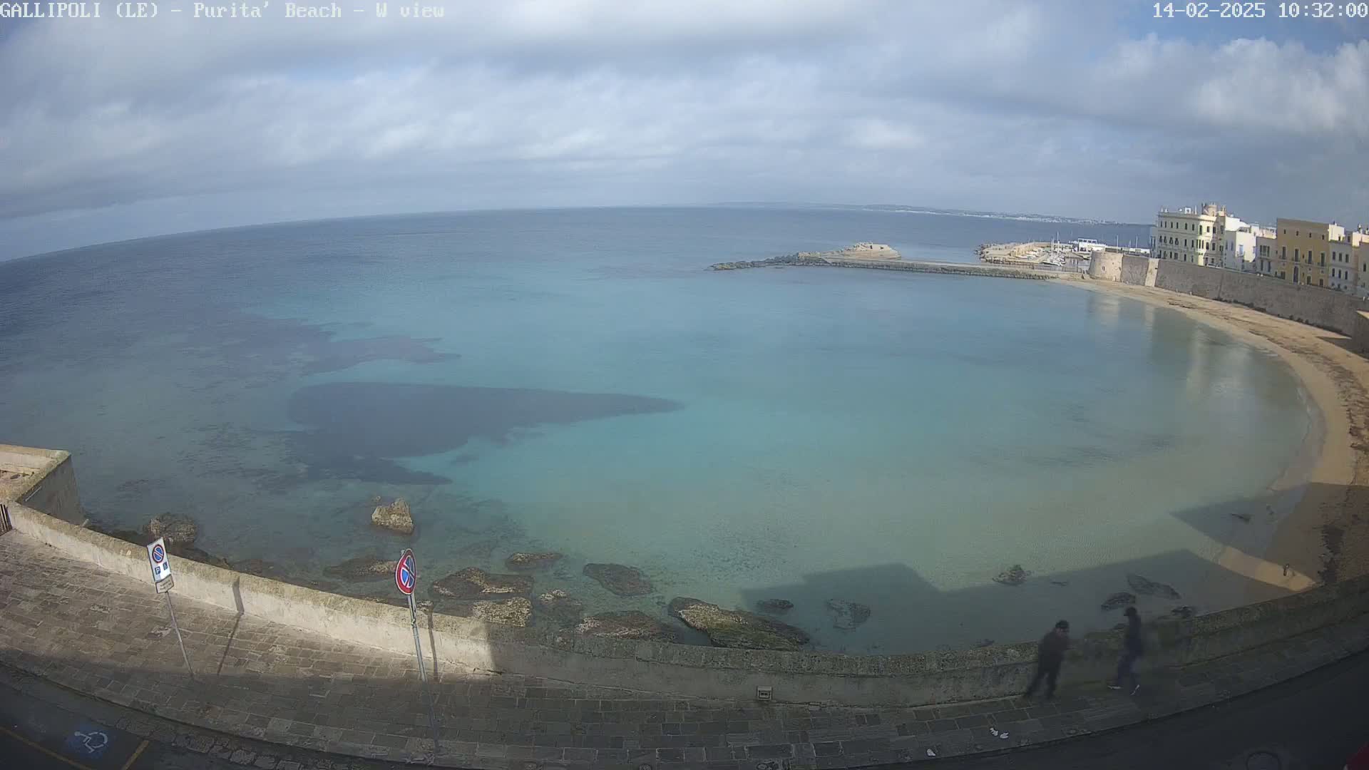 A partly cloudy day overlooks a calm, clear turquoise bay with a sandy beach and stone buildings along the shoreline.
