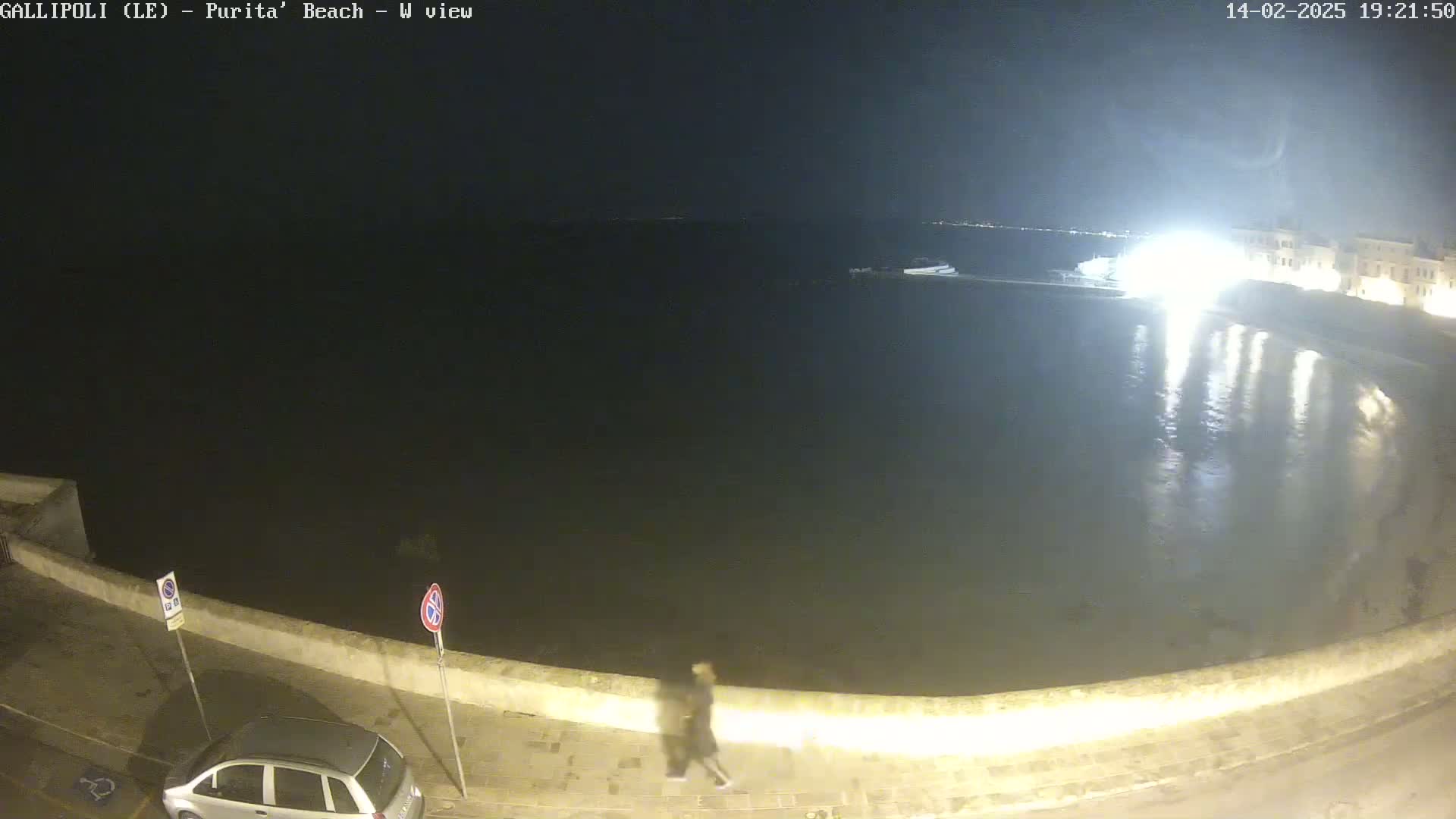 A nighttime view of a calm body of water next to a low wall, with a person walking along the wall and a car parked nearby.