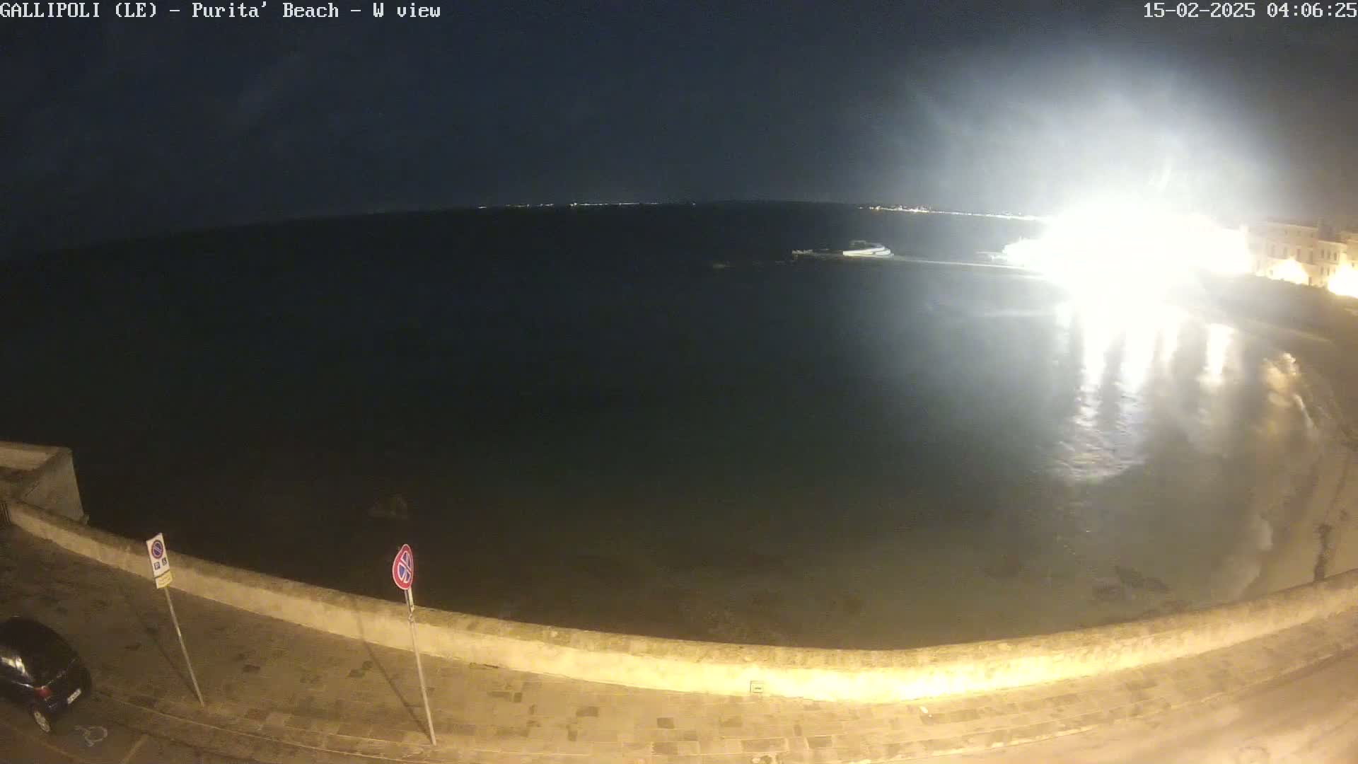 A nighttime, high-angle view shows a dark body of water next to a stone wall and paved area, with bright lights reflecting on the water's surface.